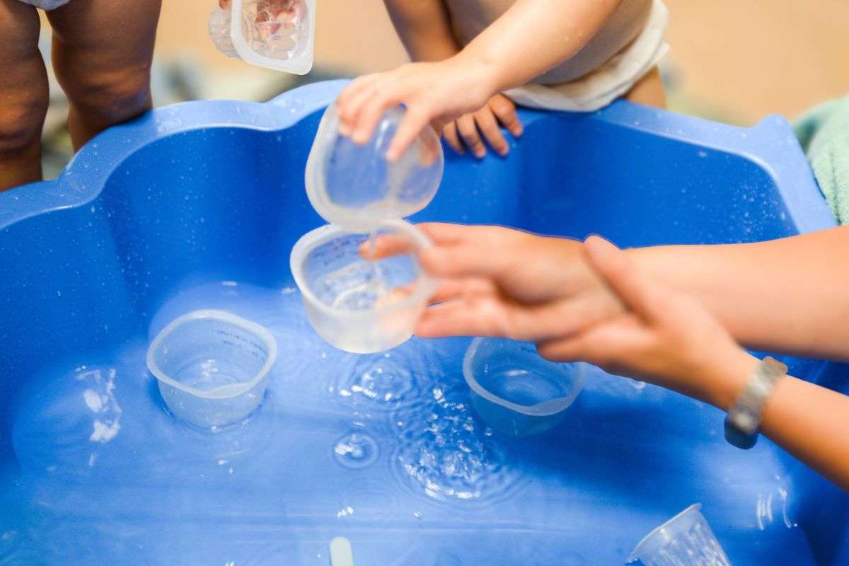 Des enfants en bas âge jouent avec de l’eau et des gobelets dans un bac bleu, sous la surveillance d’un adulte.