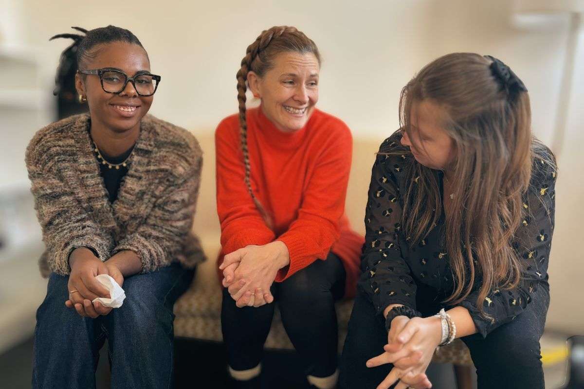 Une professionnelle de la crèche souriante, vêtue d'une chemise blanche, interagit avec une mère et son jeune enfant.