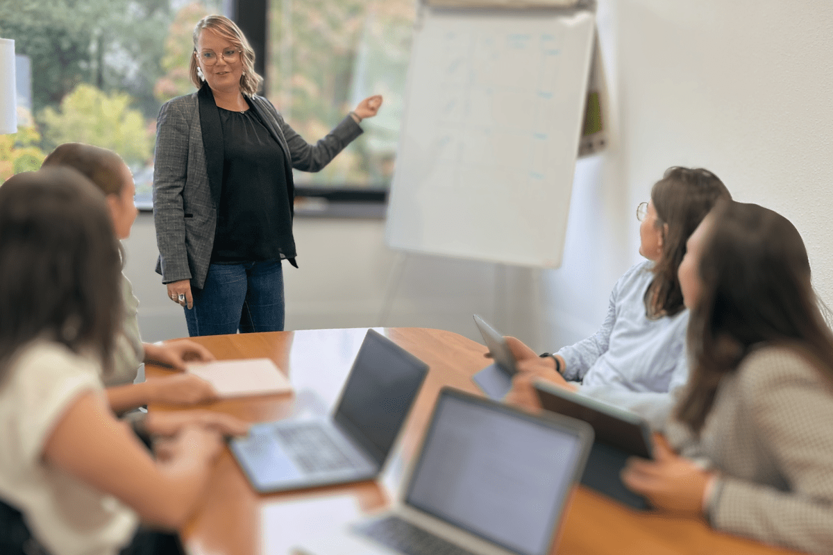 Plan sur trois femmes, membres de l'équipe de la crèche, assises autour d'une table en bois et travaillant ensemble sur des ordinateurs portables. La femme à gauche, vêtue d'une veste de costume grise, pointe du doigt l'écran d'un ordinateur. Les deux autres femmes regardent l'écran, engagées dans une discussion.